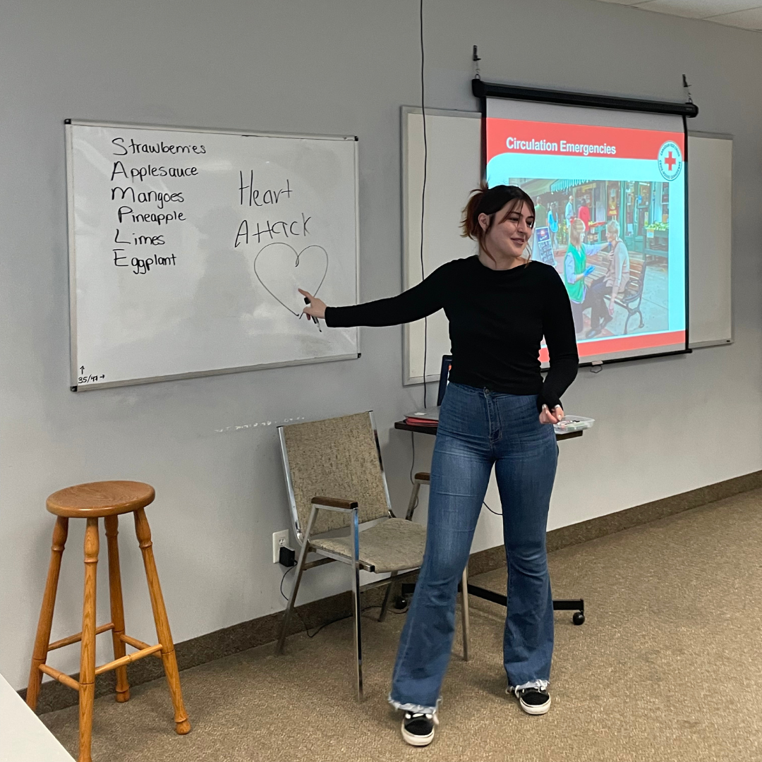 A competent and approachable female first aid instructor leading a course. She stands confidently in front of the class, demonstrating using a whiteboard. Her engaging presence fosters a positive and focused learning environment. Participants are seated attentively, listening and taking notes. The instructor is well-prepared with visual aids and a clear presentation style. The atmosphere reflects professionalism and a commitment to learning essential first aid skills.