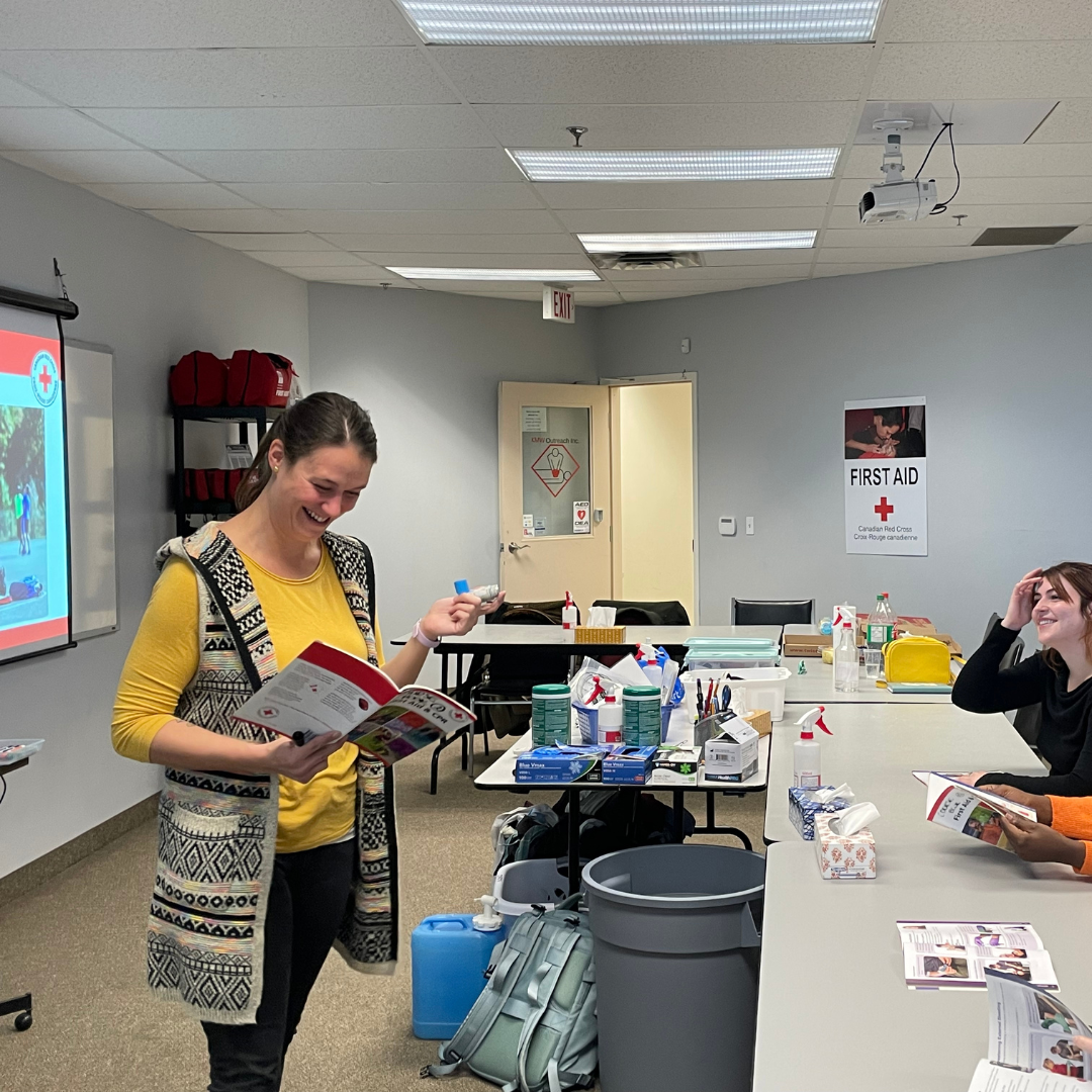 "A competent and approachable female first aid instructor leading a course. She stands confidently in front of the class, demonstrating life-saving techniques with an asthma inhaler. Her engaging presence fosters a positive and focused learning environment. Participants are seated attentively, listening and taking notes. The instructor is well-prepared with visual aids and a clear presentation style. The atmosphere reflects professionalism and a commitment to learning essential first aid skills.