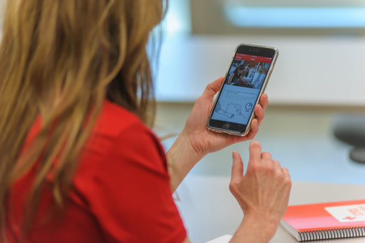 A woman engaged in a first aid course on her mobile phone. She is focused on the screen, participating in an online learning platform. This image reflects a modern approach to first aid education, with the woman using technology for training and skill development.