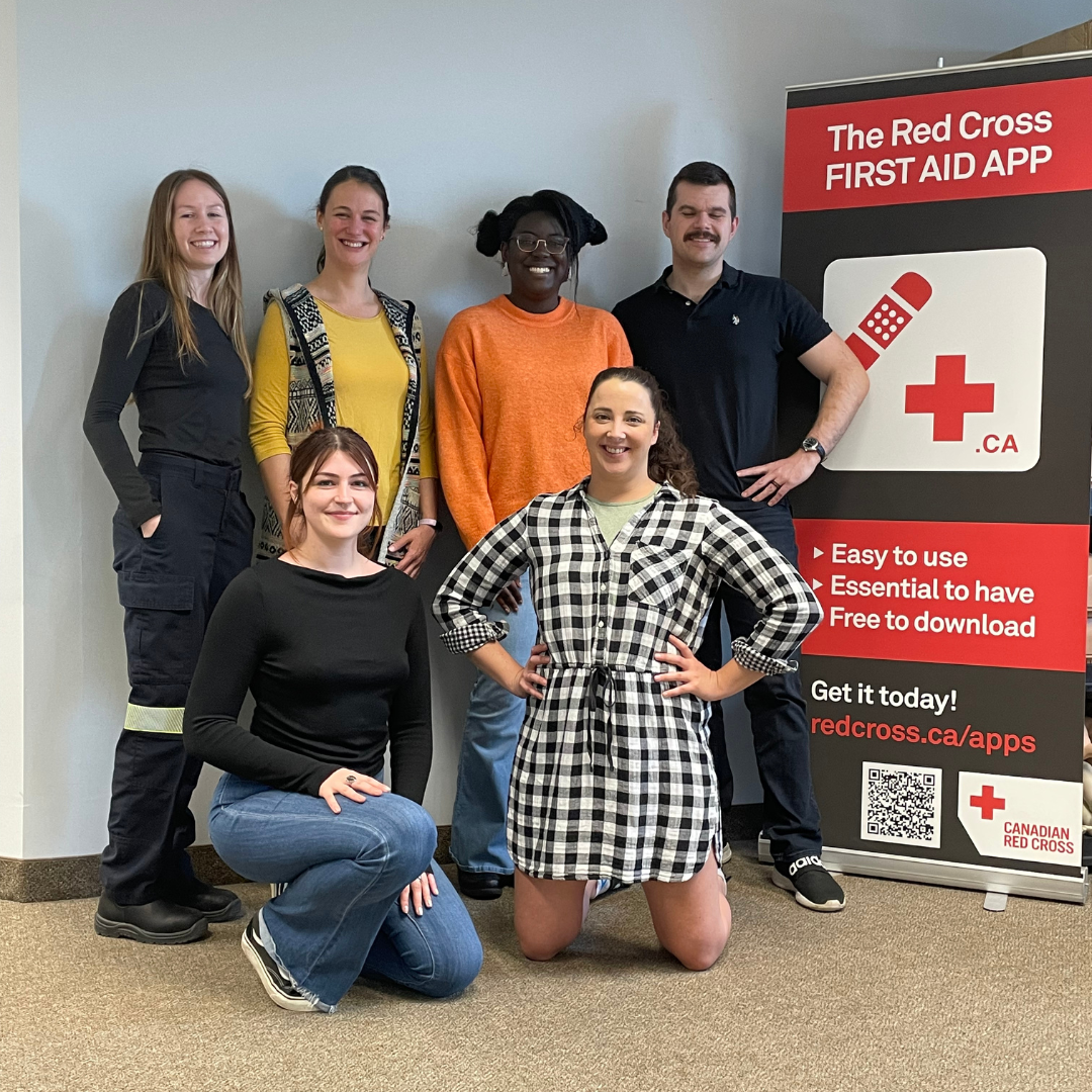Team photo of KMW Outreach Inc. employees, gathered in a group. Smiling faces of diverse individuals representing various departments and roles within the company. The atmosphere is positive and collaborative, with employees standing or sitting together. The background features the Canadian Red Cross logo and a vibrant office space. This photo captures the sense of unity and teamwork within the company